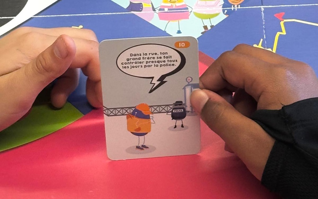 Zoom sur la carte Diskri’Minis montrant un exemple de situation sur les contrôles policiers répétés et la discrimination, tenue par un élève.