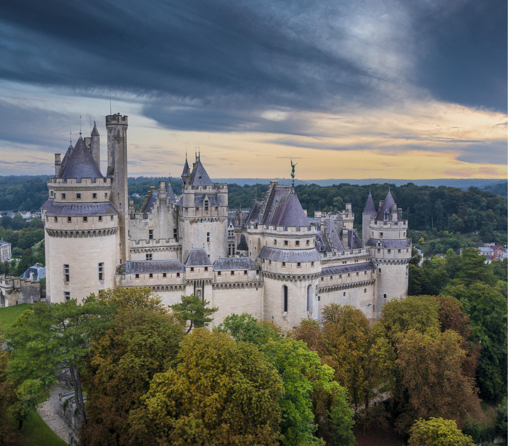 Vue aérienne du château médiéval de Pierrefonds entouré de forêt sous un ciel nuageux.