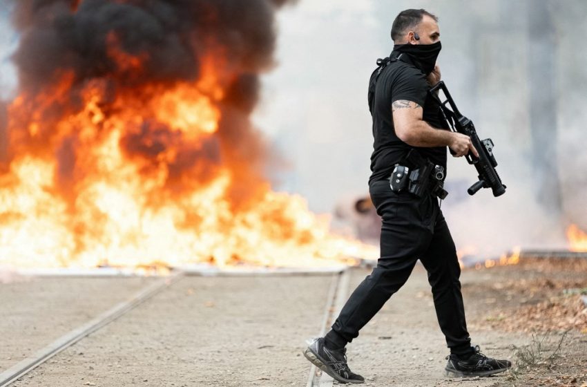 Policier portant un lanceur LBD-40 devant des poubelles en feu lors d’une manifestation lycéenne à Strasbourg.